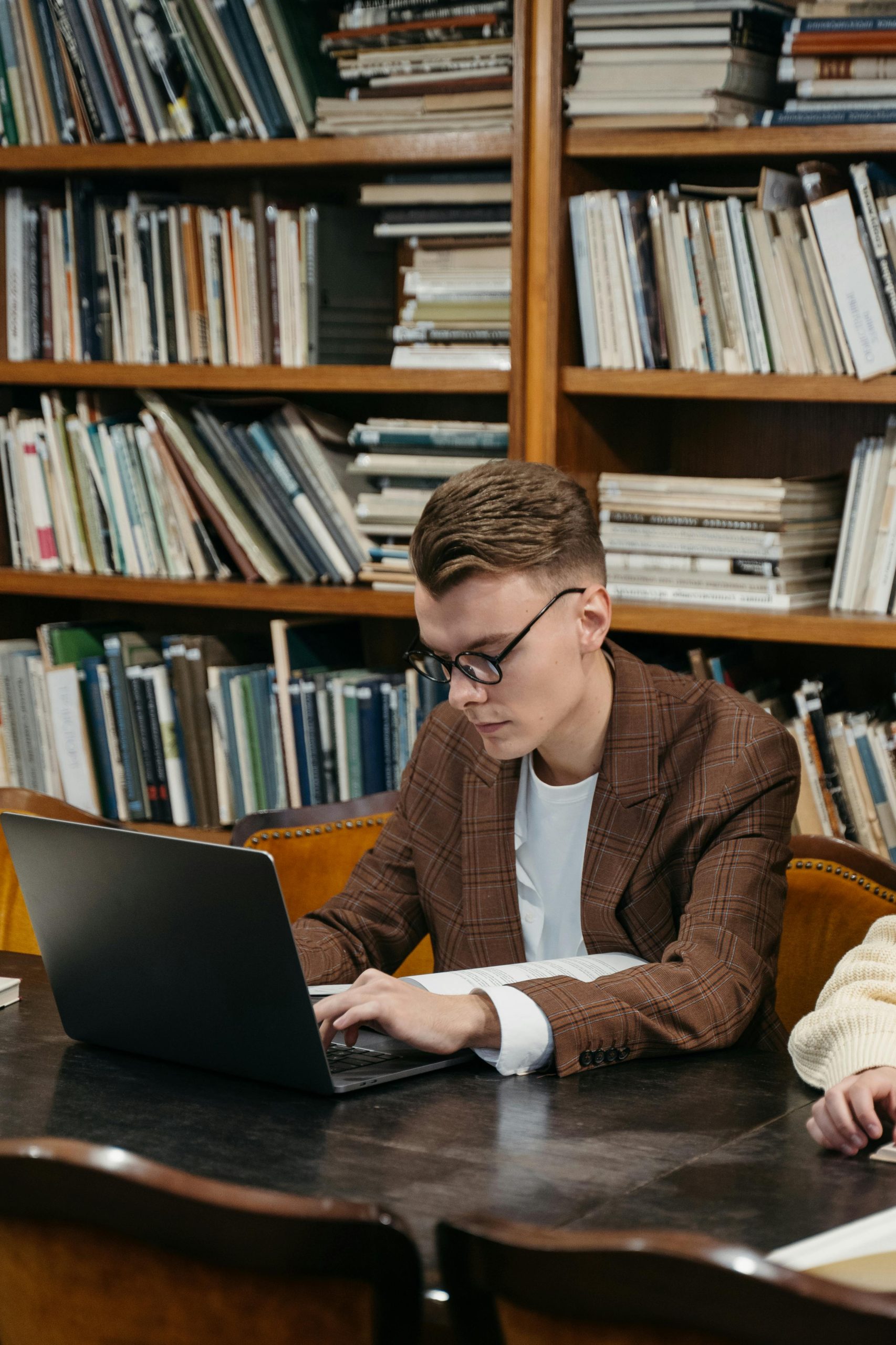 A focused young man working on a laptop surrounded by books in a library setting.